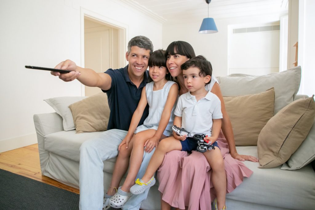 happy parent couple with two kids watching tv sitting on couch in living room and using remote control